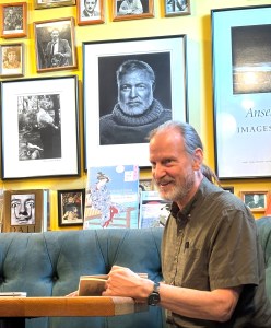 White male with graying beard and blond pony tail reading poetry. Pictures of Ernest Hemmingway and other writers/artist in the background.